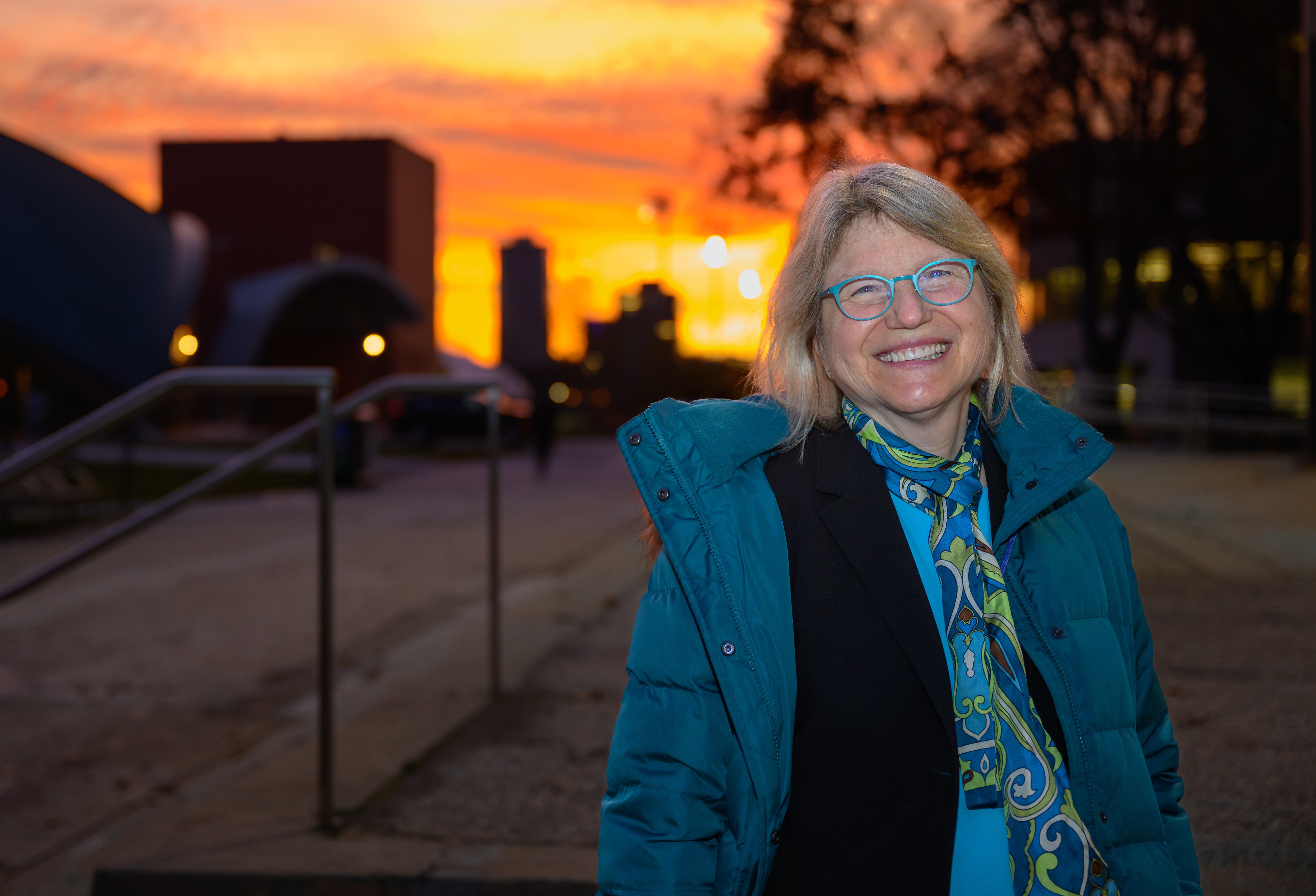 President Kornbluth poses smiling with Kresge Auditorium, the Linde Music Building, and an orange sunset in the background.