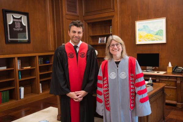 Mark Rober (left) dressed in a black robe stands next to President Kornbluth (right) dressed in a gray and red robe in her office posing together for commencement photos.