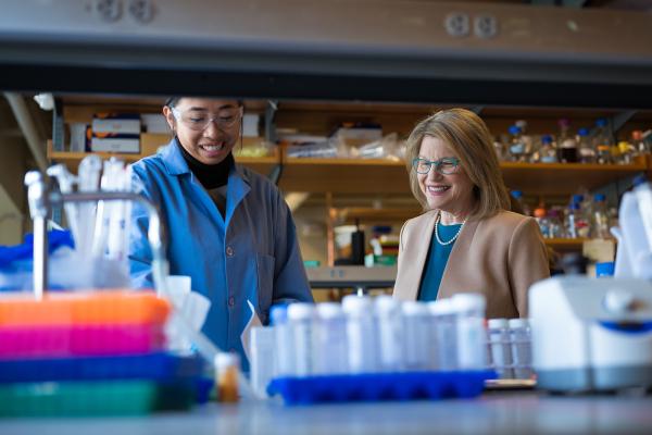President Kornbluth stands next to student Victoria Yang as they both observe the experiment and lab equipment in the foreground. 
