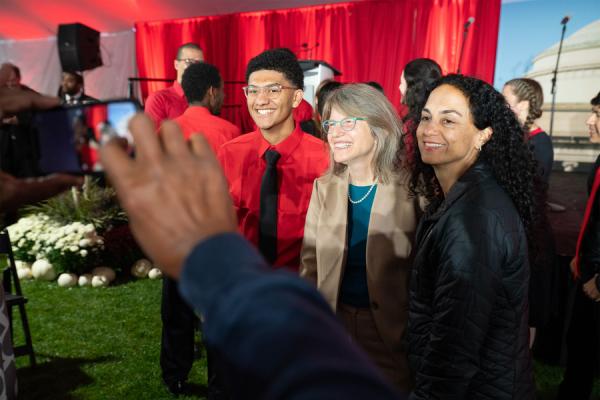 Two people stand on either side of Sally Kornbluth. All three are smiling as another person takes a photo of them.