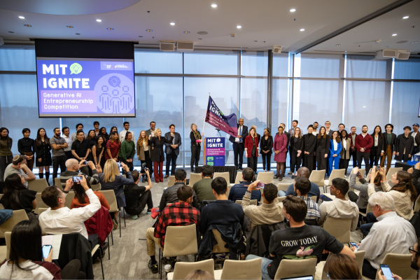 President Kornbluth stands next to a podium and holds the flag for MIT Ignite: Generative AI Entrepreneurship Competition in front of a crowd.