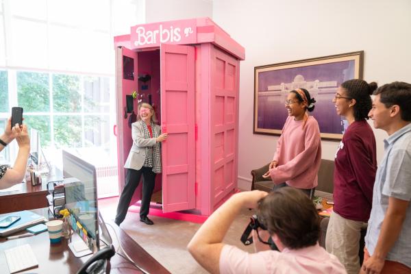 President Kornbluth laughs and wears pink sunglasses as she steps out of the pink Barbis phone booth while students laugh and observe.